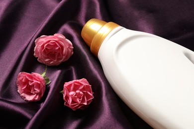 Photo of Bottle of fabric softener and flowers on purple cloth, closeup