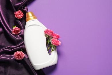 Photo of Bottle of fabric softener, flowers and color cloth on purple background, flat lay. Space for text