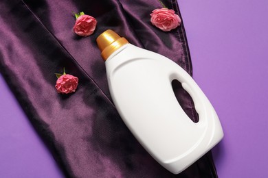 Photo of Bottle of fabric softener, flowers and color cloth on purple background, flat lay