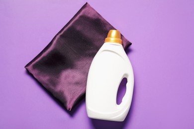 Photo of Bottle of fabric softener and color cloth on purple background, top view