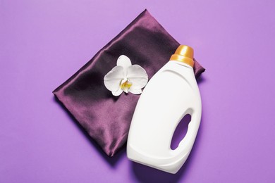 Photo of Bottle of fabric softener, orchid flower and color cloth on purple background, top view