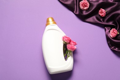 Photo of Bottle of fabric softener, flowers and color cloth on purple background, flat lay