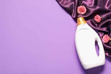 Photo of Bottle of fabric softener, flowers and color cloth on purple background, flat lay. Space for text