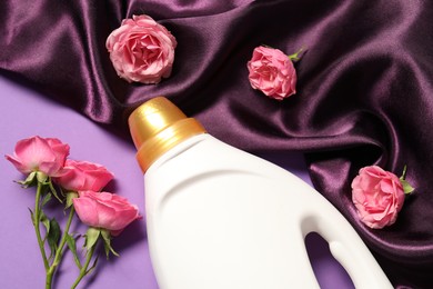 Photo of Bottle of fabric softener, flowers and color cloth on purple background, flat lay