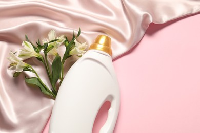 Photo of Bottle of fabric softener, flowers and beige cloth on pink background, flat lay. Space for text