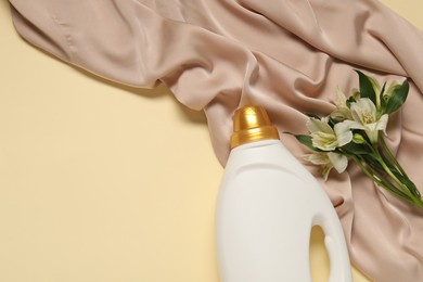 Photo of Bottle of fabric softener, flowers and color cloth on beige background, flat lay. Space for text