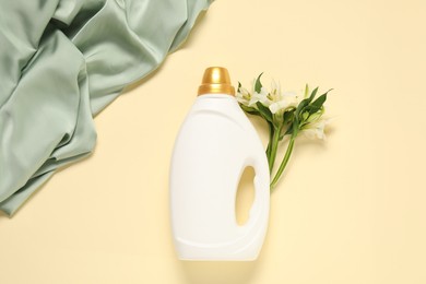 Photo of Bottle of fabric softener, flowers and green cloth on beige background, flat lay
