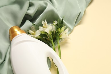Photo of Bottle of fabric softener, flowers and green cloth on beige background, above view. Space for text