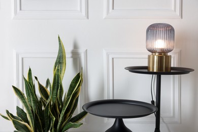 Photo of Lamp, stylish black coffee tables and houseplant near white wall indoors