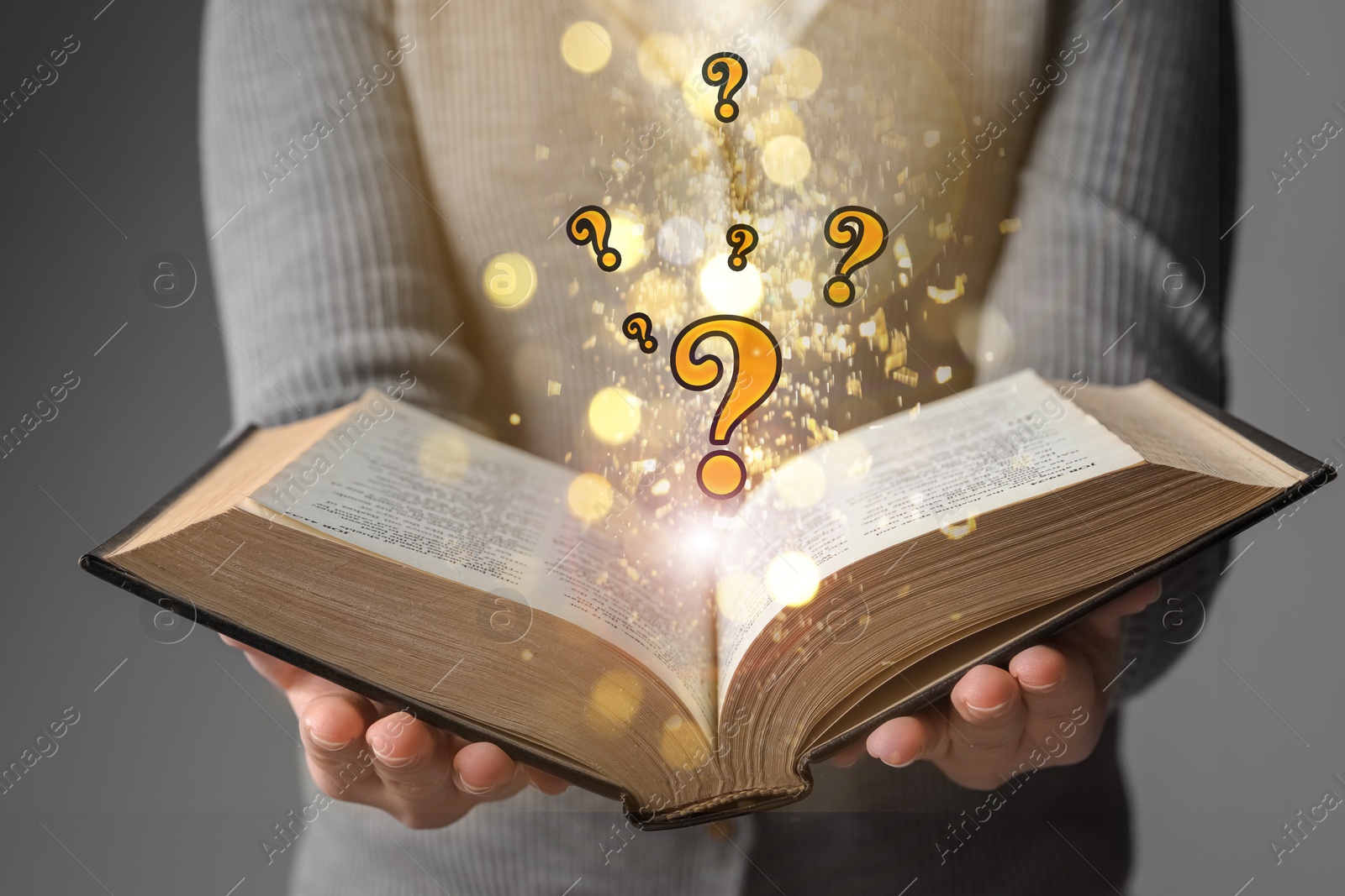 Woman holding open book against grey background, closeup. Question marks and magic lights above pages Image of Woman holding open book against grey background, closeup. Question marks and magic lights above pages