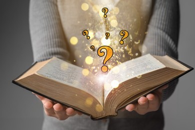 Image of Woman holding open book against grey background, closeup. Question marks and magic lights above pages