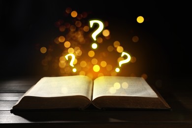 Image of Question marks and magic lights flying out of open book on table against black background