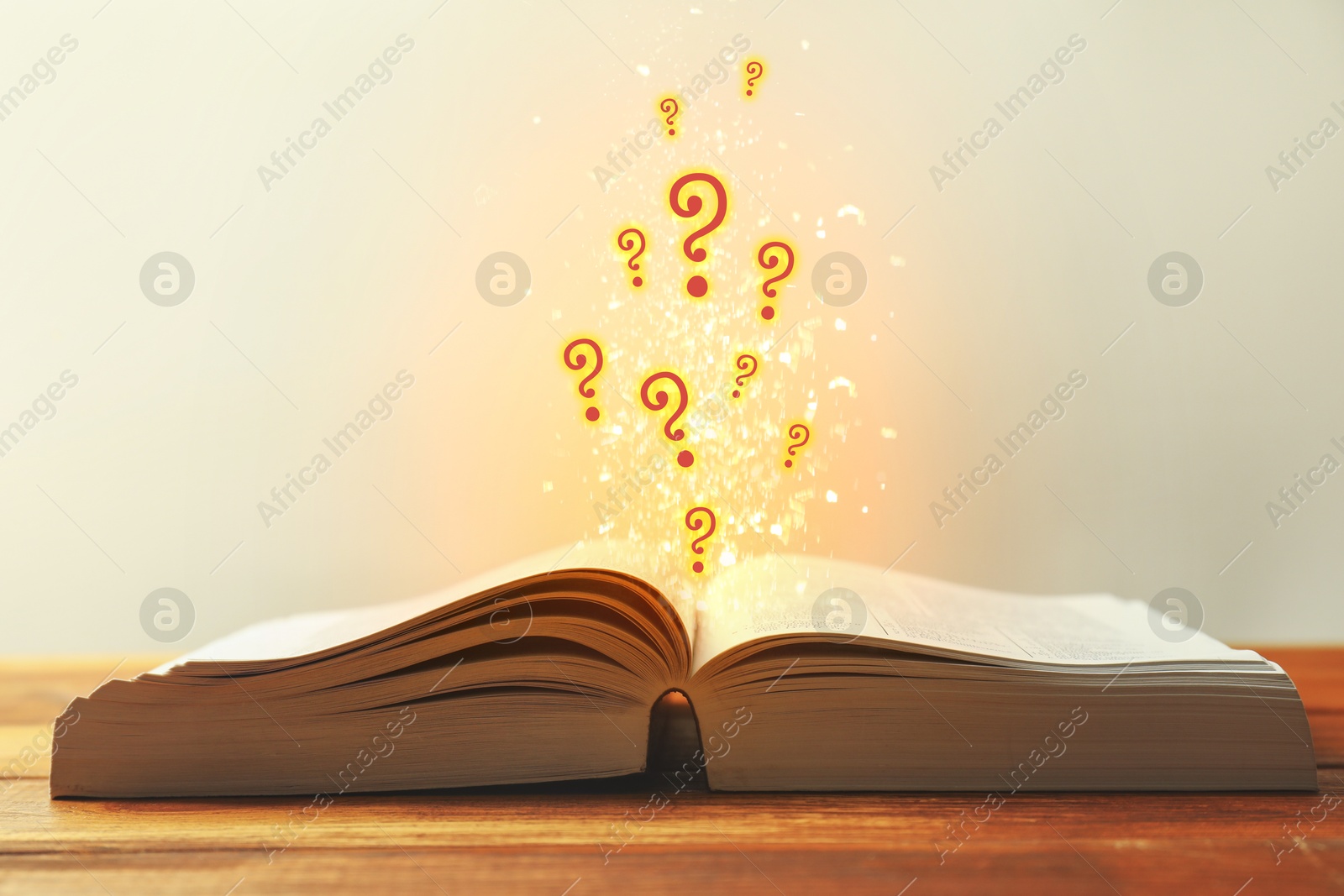 Question marks and lights flying out of open book on table against light background Image of Question marks and lights flying out of open book on table against light background