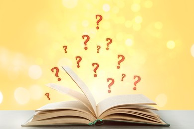 Image of Question marks above open book on table against yellow background with blurred lights