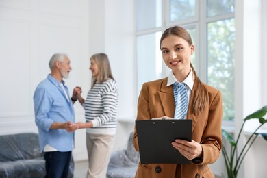 Photo of Smiling real estate agent with clipboard and couple at home, selective focus