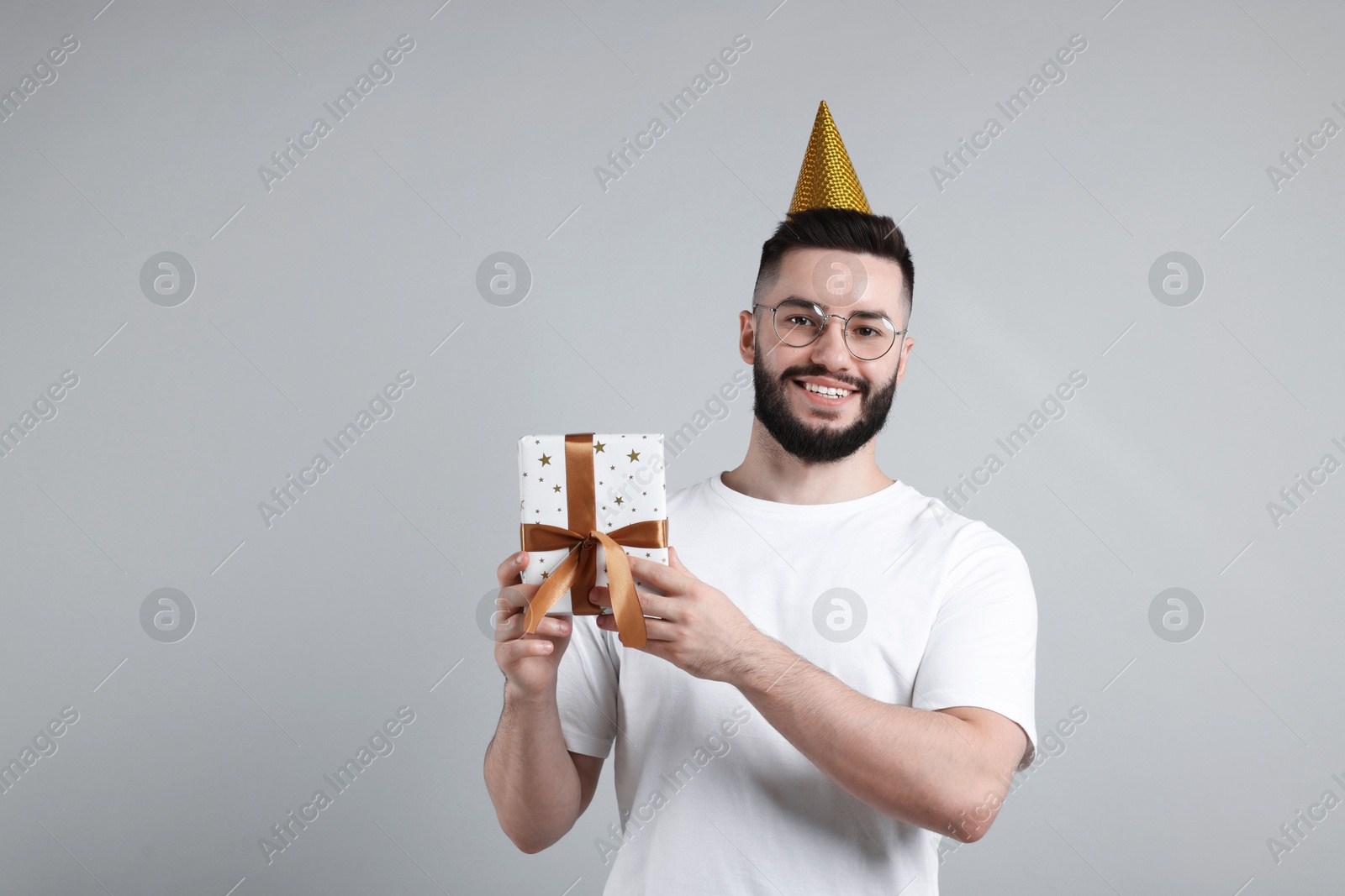 Happy Birthday. Handsome man with party hat and gift box on grey background, space for text Photo of Happy Birthday. Handsome man with party hat and gift box on grey background, space for text