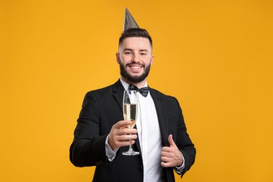 Photo of Happy Birthday. Handsome man with party hat and champagne showing thumbs up on orange background
