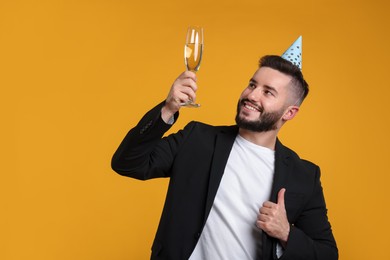 Photo of Happy Birthday. Handsome man with party hat and champagne showing thumbs up on orange background, space for text