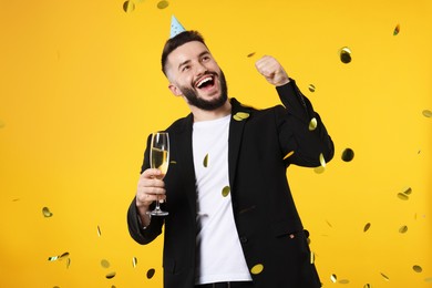 Photo of Happy Birthday. Handsome man with party hat and glass of champagne under confetti shower on orange background