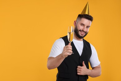 Photo of Happy Birthday. Handsome man with party hat and glass of champagne showing thumbs up on orange background, space for text