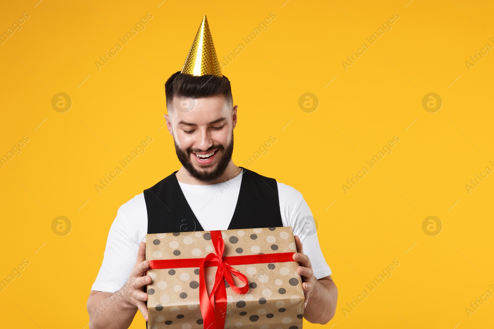 Happy Birthday. Handsome man with party hat and gift box on orange background, space for text Photo of Happy Birthday. Handsome man with party hat and gift box on orange background, space for text
