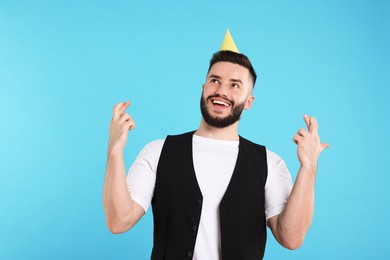 Photo of Happy Birthday. Handsome man with party hat crossing fingers while making wish on light blue background
