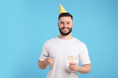 Photo of Happy Birthday. Handsome man with party hat, blower and glass of champagne on light blue background
