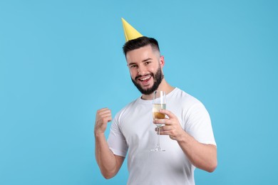 Photo of Happy Birthday. Handsome man with party hat and glass of champagne on light blue background, space for text