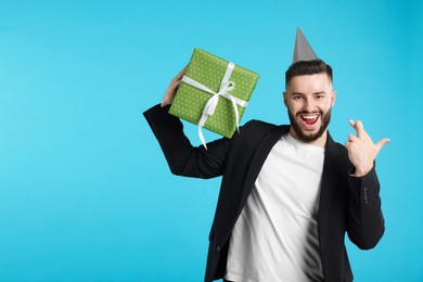 Photo of Happy Birthday. Handsome man with party hat and gift box crossing fingers while making wish on light blue background, space for text