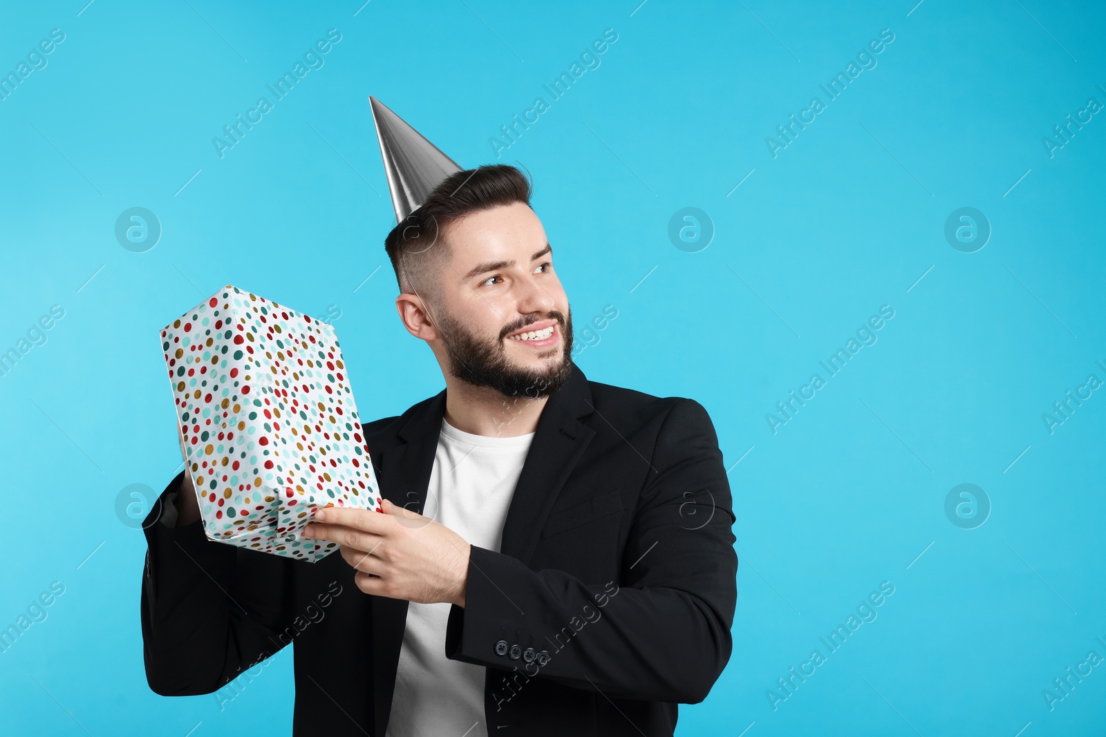 Happy Birthday. Handsome man with party hat and gift box on light blue background, space for text Photo of Happy Birthday. Handsome man with party hat and gift box on light blue background, space for text