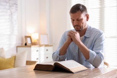 Photo of Man with Bible praying at table indoors. Space for text