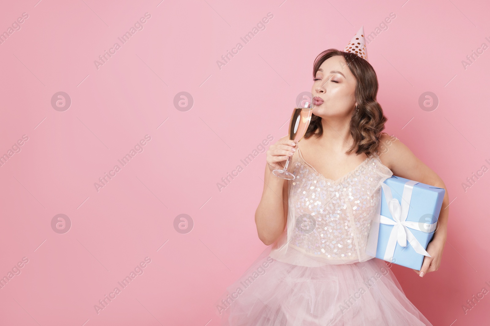 Birthday celebration. Woman with glass of sparkling wine and gift on pink background. Space for text Photo of Birthday celebration. Woman with glass of sparkling wine and gift on pink background. Space for text