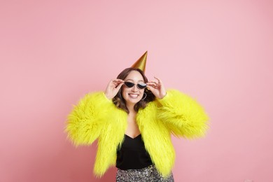 Photo of Birthday celebration. Happy woman with party cone and sunglasses on pink background