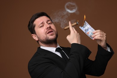 Photo of Greedy banker lighting cigar with dollar banknote on brown background