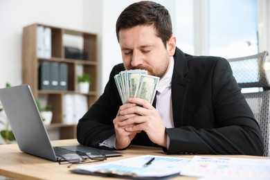 Photo of Greedy banker sniffing dollar banknotes at table in office