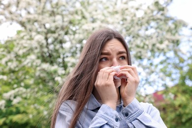Photo of Woman with paper tissue suffering from seasonal pollen allergy near blossoming tree on spring day