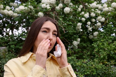 Photo of Woman with paper tissue suffering from seasonal pollen allergy near blossoming shrub on spring day, space for text