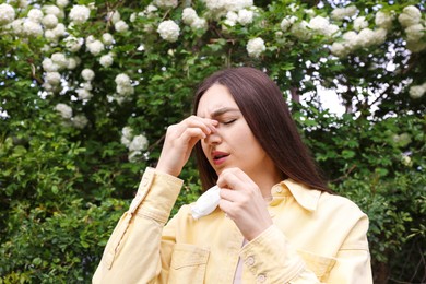 Photo of Woman with paper tissue suffering from seasonal pollen allergy near blossoming shrub on spring day