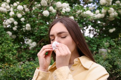 Photo of Woman with paper tissue suffering from seasonal pollen allergy near blossoming shrub on spring day