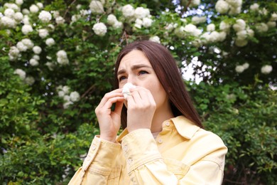 Photo of Woman with paper tissue suffering from seasonal pollen allergy near blossoming shrub on spring day