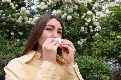 Photo of Woman with paper tissue suffering from seasonal pollen allergy near blossoming shrub on spring day, space for text
