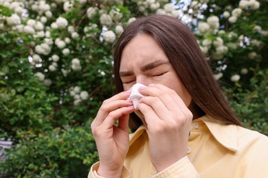 Photo of Woman with paper tissue suffering from seasonal pollen allergy near blossoming shrub on spring day, space for text
