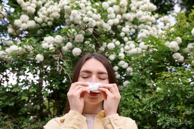 Photo of Woman with paper tissue suffering from seasonal pollen allergy near blossoming shrub on spring day, low angle view