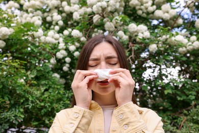 Photo of Woman with paper tissue suffering from seasonal pollen allergy near blossoming shrub on spring day