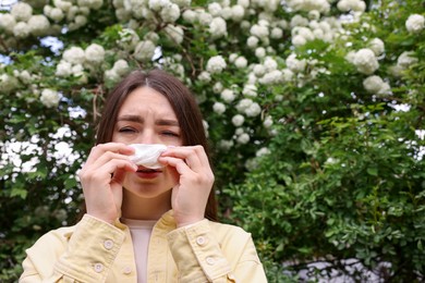 Photo of Woman with paper tissue suffering from seasonal pollen allergy near blossoming shrub on spring day, space for text