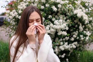 Photo of Woman with paper tissue suffering from seasonal pollen allergy near blossoming shrub on spring day, space for text