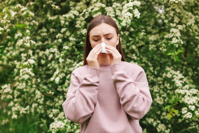 Photo of Woman with paper tissue suffering from seasonal pollen allergy near blossoming shrub on spring day