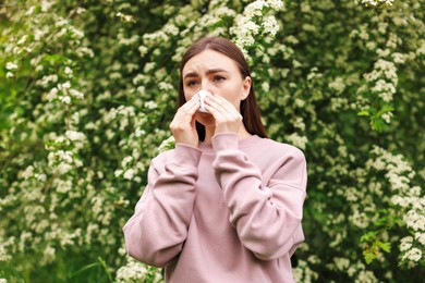 Photo of Woman with paper tissue suffering from seasonal pollen allergy near blossoming shrub on spring day