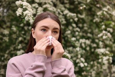 Photo of Woman with paper tissue suffering from seasonal pollen allergy near blossoming shrub on spring day, space for text