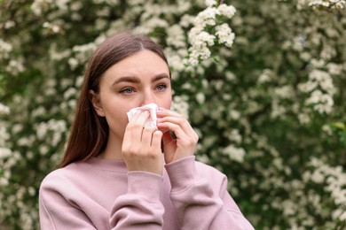 Photo of Woman with paper tissue suffering from seasonal pollen allergy near blossoming shrub on spring day, space for text
